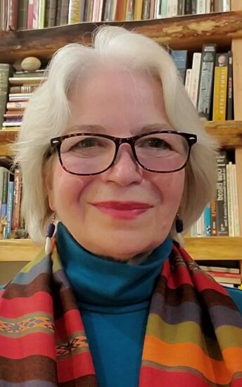 Image of a white woman with white hair wearing a colorful scarf, blue shirt, and tortoise shell spectacles