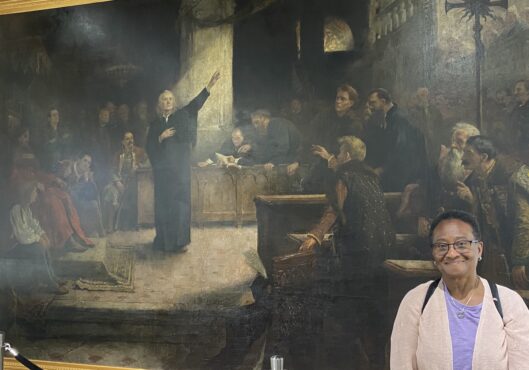 Image of a black woman wearing a purple shirt and pink cardigan in front of a large painting of the Edict of Torta