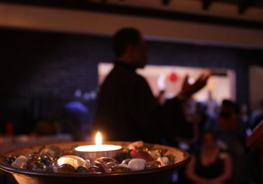 Image of a lit chalice in the foreground with a black minister preaching in the background