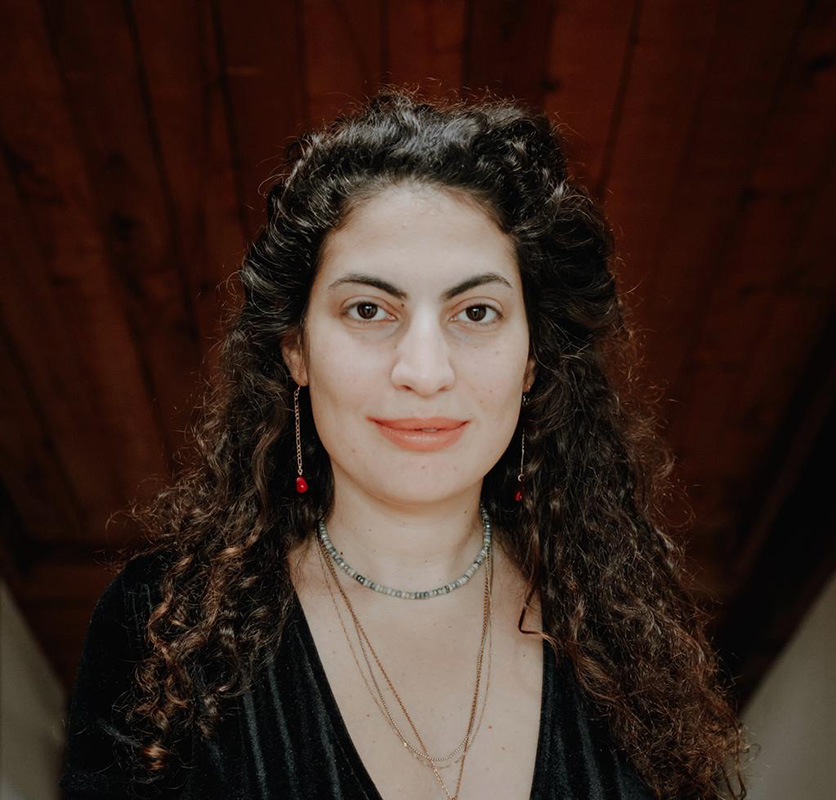 Image of an Arab Jewish woman with jewelry and a gray and black striped top set in front of a dark wooden background