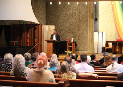 A person in a black robe speaks at a podium in a church with an audience seated in pews. The church features a projector screen, a wooden backdrop, and a colorful fabric arc on one wall.