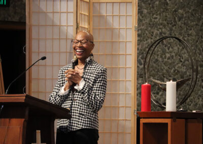 A person wearing glasses and a black-and-white checkered blazer stands happily behind a lectern, hands clasped together. There is a microphone in front of them and a screen divider in the background. To their right is a wooden stand with a metal chalice, red and white candles.