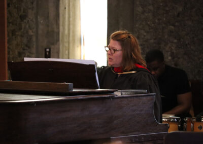 A person with glasses and shoulder-length hair, in a robe with a red collar, is passionately singing while playing the piano. The dimly lit room with stone walls adds to the ambiance of this enchanting event.