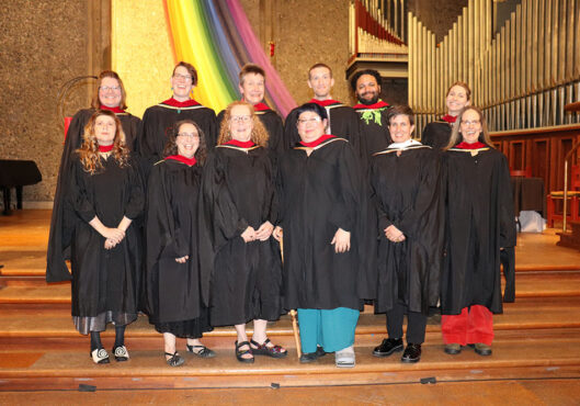 A group of eleven people wearing academic robes and caps smile for a group photo in front of a church altar during an event at the Unitarian Universalist Church of Berkeley. Behind them is a large organ and a rainbow banner draped along the wall, suggesting a celebratory or graduation event.