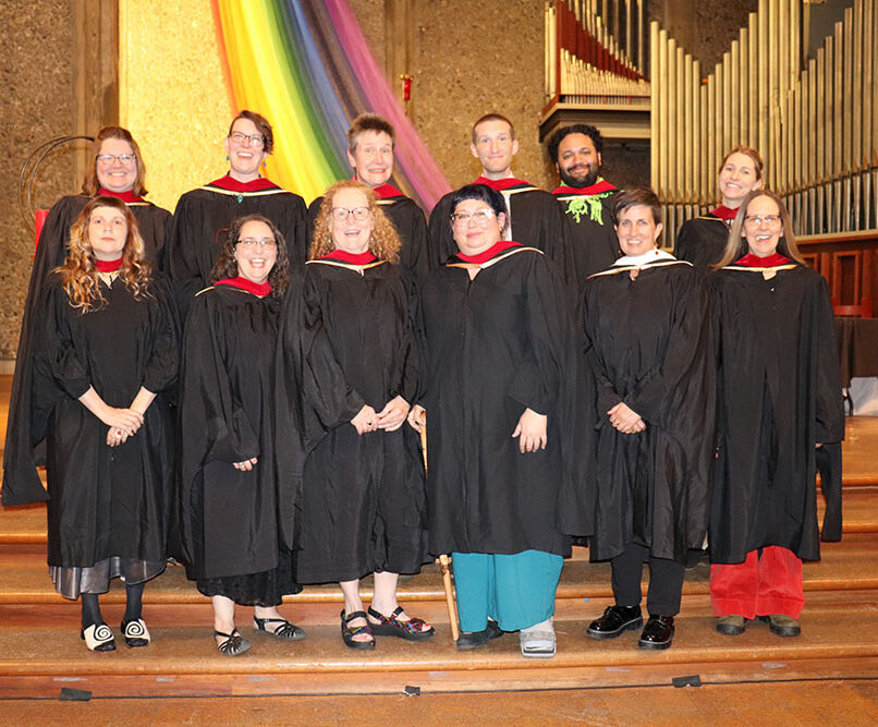 A group of eleven people wearing academic robes and caps smile for a group photo in front of a church altar during an event at the Unitarian Universalist Church of Berkeley. Behind them is a large organ and a rainbow banner draped along the wall, suggesting a celebratory or graduation event.
