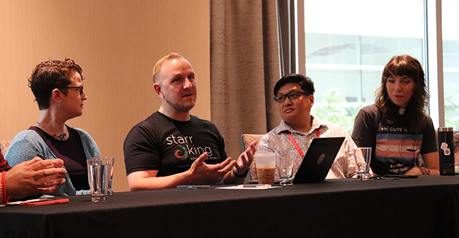 Four people sit at a conference table engaged in discussion. One person wearing a Starr King School for the Ministry shirt is speaking and gesturing with his hands. They have laptops, coffee, and water glasses in front of them. A large window in the background lets in natural light.