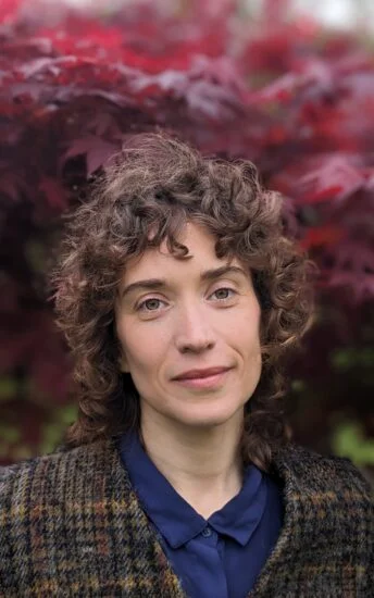 A calm Rabbi with curly hair stands in front of a tree with vibrant red leaves. Wearing a checked coat over a blue shirt, Ora Nitkin-Kaner commands attention as the background softly blurs around them.