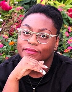Meet Our featured individual: A person with short hair, wearing glasses and a nose ring, is looking at the camera with a calm expression. Their hand is slightly touching their chin. The background features vibrant flowers and greenery from the St. Lawrence Scholars garden, 2023-2024.