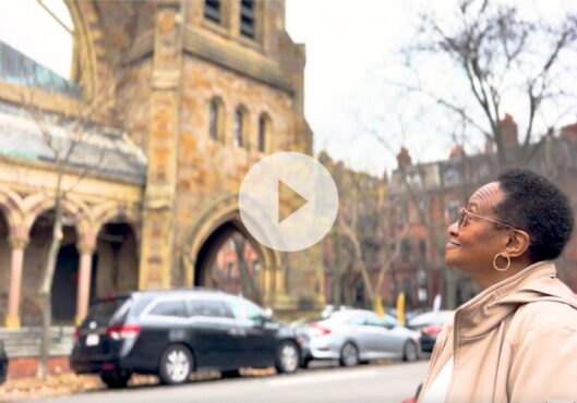 An elderly woman with glasses and short hair stands on a sidewalk, gazing up at a historic stone building with arched windows. The building's intricate architecture is prominent, and parked cars line the street. The scene is peaceful, with bare trees in the background and an air of anticipation for the President’s special announcement.