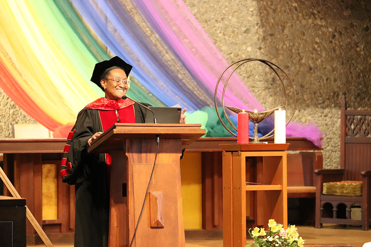 A speaker in academic regalia, including a black cap and red gown, stands at a podium while giving a speech. Behind them is a colorful rainbow fabric draped as a backdrop.