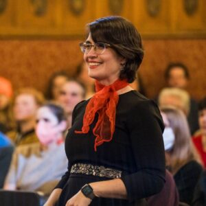 Meet Our featured individual: A person with short hair, wearing glasses and a nose ring, is looking at the camera with a calm expression. Their hand is slightly touching their chin. The background features vibrant flowers and greenery from the St. Lawrence Scholars garden, 2023-2024.